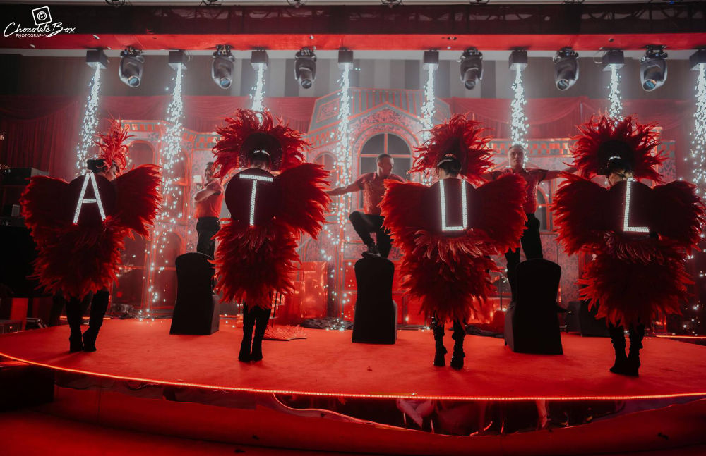 Performers in red feather costumes on stage with festive lights, spelling out ATUL in dramatic fashion.