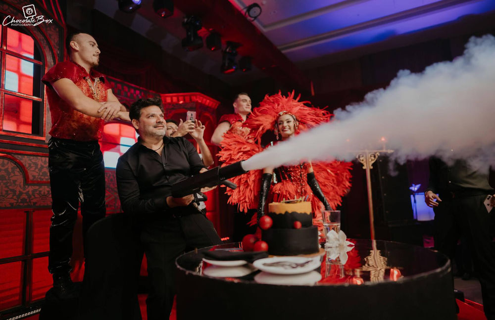 Man joyfully using a smoke gun at a lively party with colorful decor and performers in vibrant costumes.