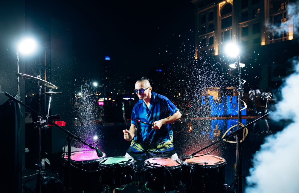 Drummer performs energetically with water splashing under colorful lights at a vibrant outdoor night event.