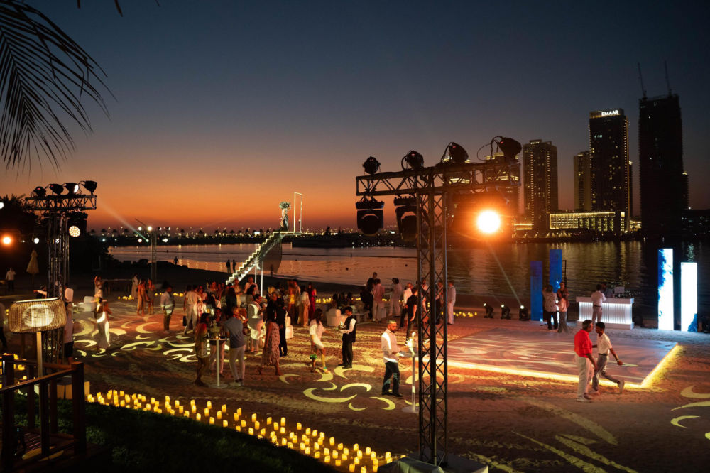 Sunset beach party with vibrant lights, people dancing, and city skyline in the background.