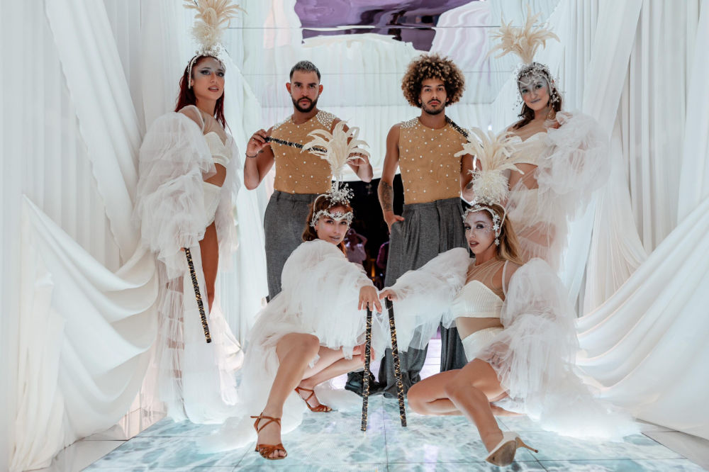 Group in elegant costumes posing on a glossy floor with white drapes and feathered headpieces.