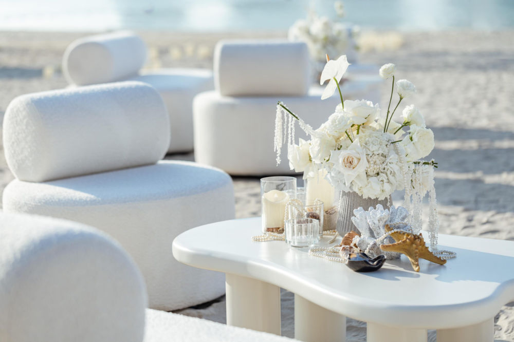 Beachfront lounge with elegant white chairs and table adorned with floral centerpiece, seashells, and candles.