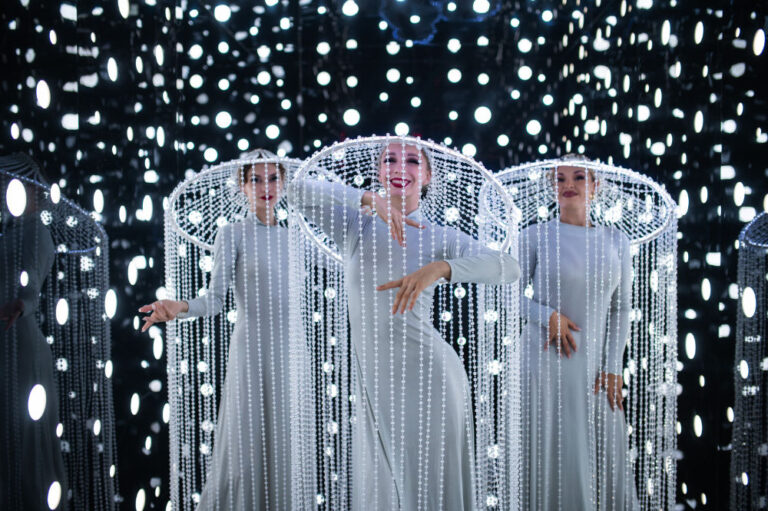 Performers in white costumes with beaded headdresses, surrounded by bright lights in a dazzling art installation.