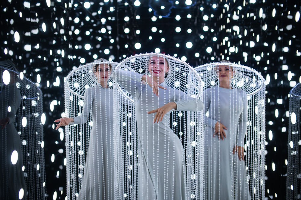 Performers in white costumes with beaded headdresses, surrounded by bright lights in a dazzling art installation.