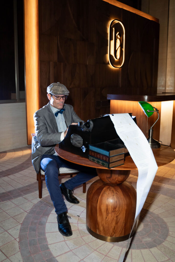 Man typing on vintage typewriter at wooden desk, long paper roll, and books nearby in stylish room.