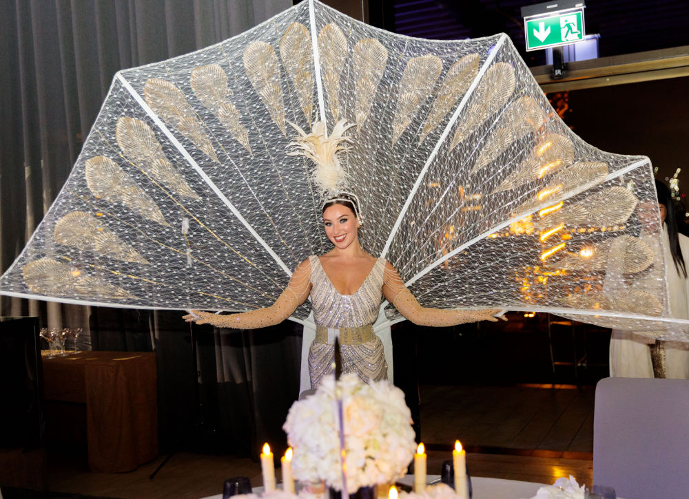 Performer in elegant costume with large, decorative wings at an event. Candles and flowers in foreground.