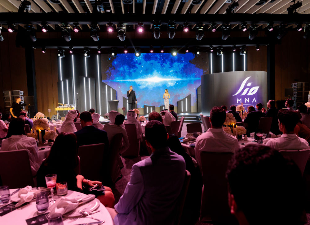 Conference event with a stage presentation, audience seated at tables, colorful lighting, and large display screen.