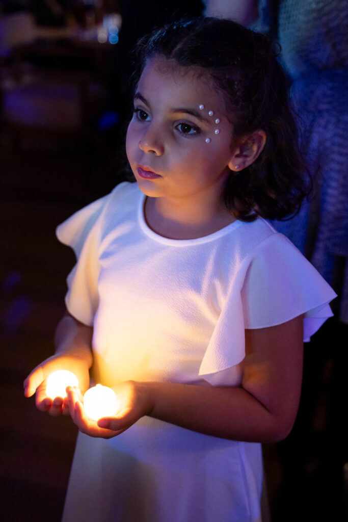 Young girl holding glowing candles, wearing white dress with jeweled face design, in dimly lit setting.