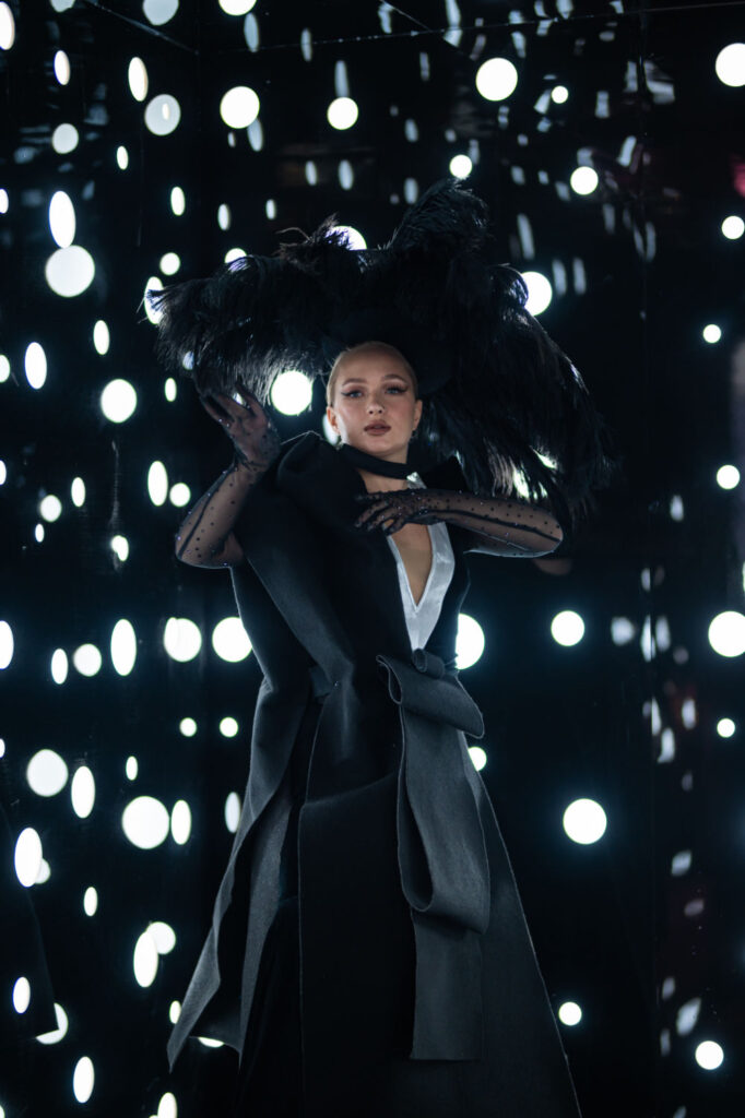 Elegant person in dark fashion with feathery hat, surrounded by glowing lights in mirrored room.
