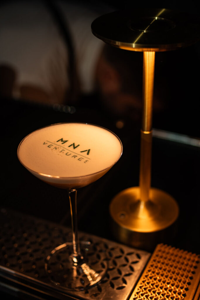 Elegant cocktail on bar, illuminated by warm light, featuring MNA Ventures logo on frothy surface.