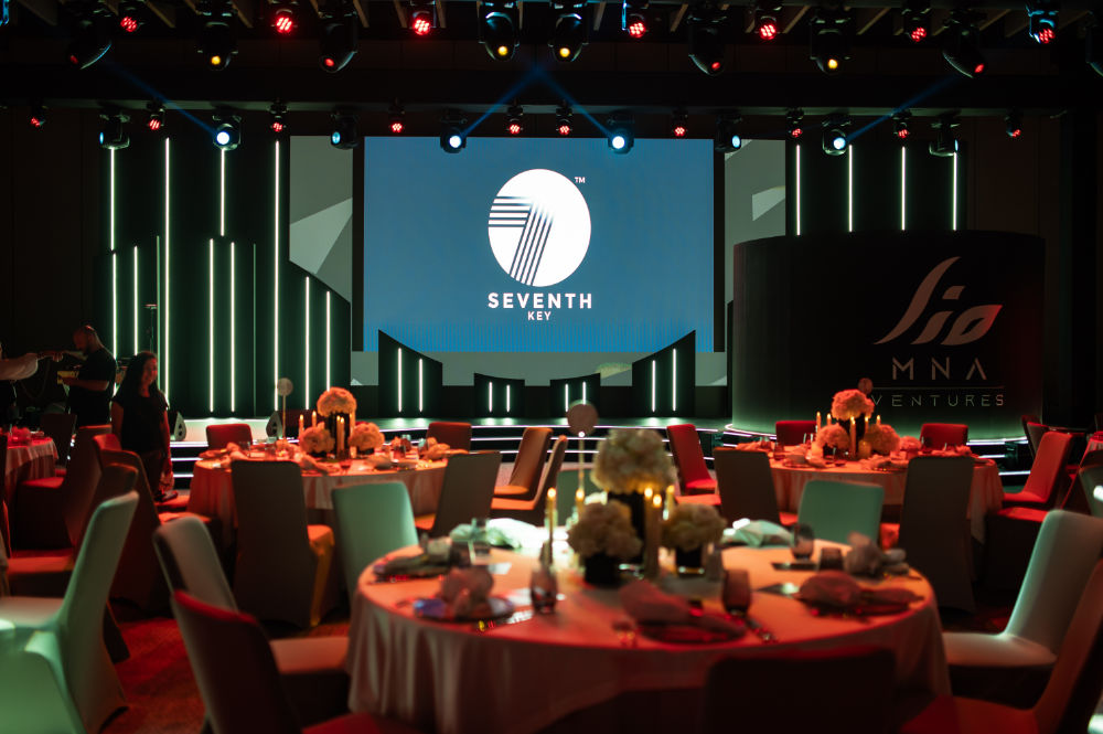 Elegant event setup with round tables and floral decor, vibrant stage lighting, and Seventh Key logo on screen.