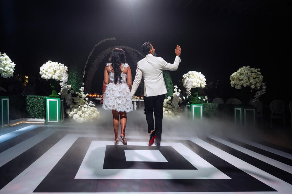 Couple in elegant attire on a floral-decorated stage, ambience lit with dramatic lights and mist.
