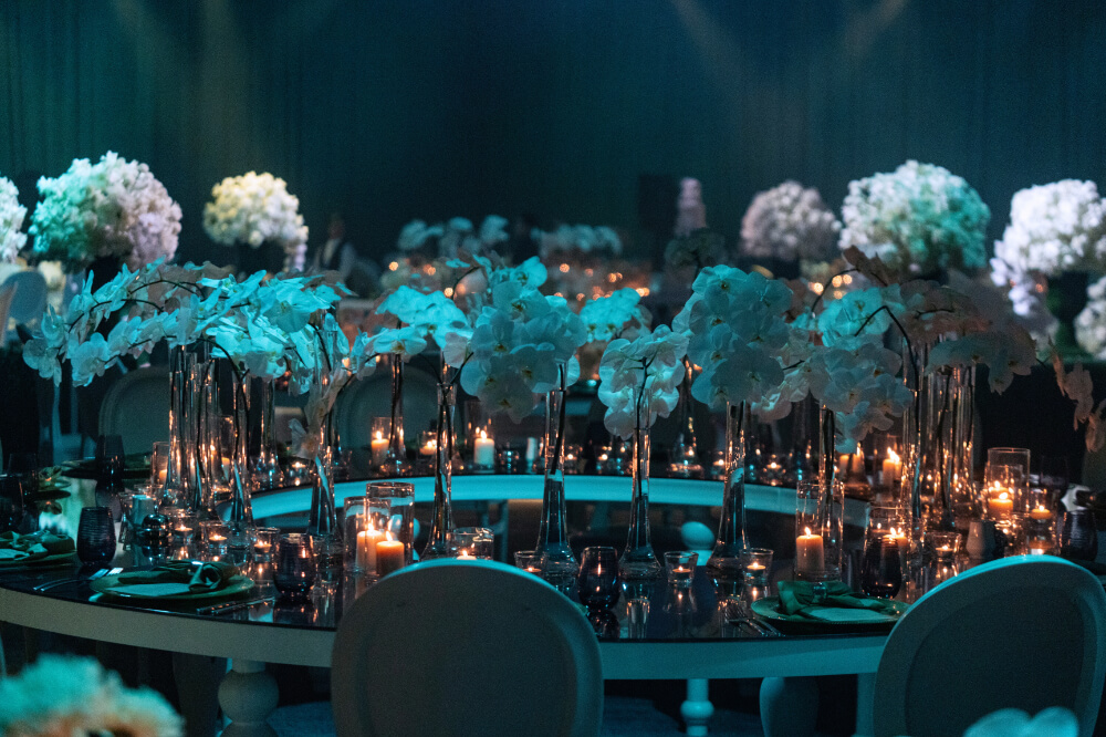 Elegant wedding table setting with white orchids, candles, and lush floral arrangements in a dimly lit banquet hall.