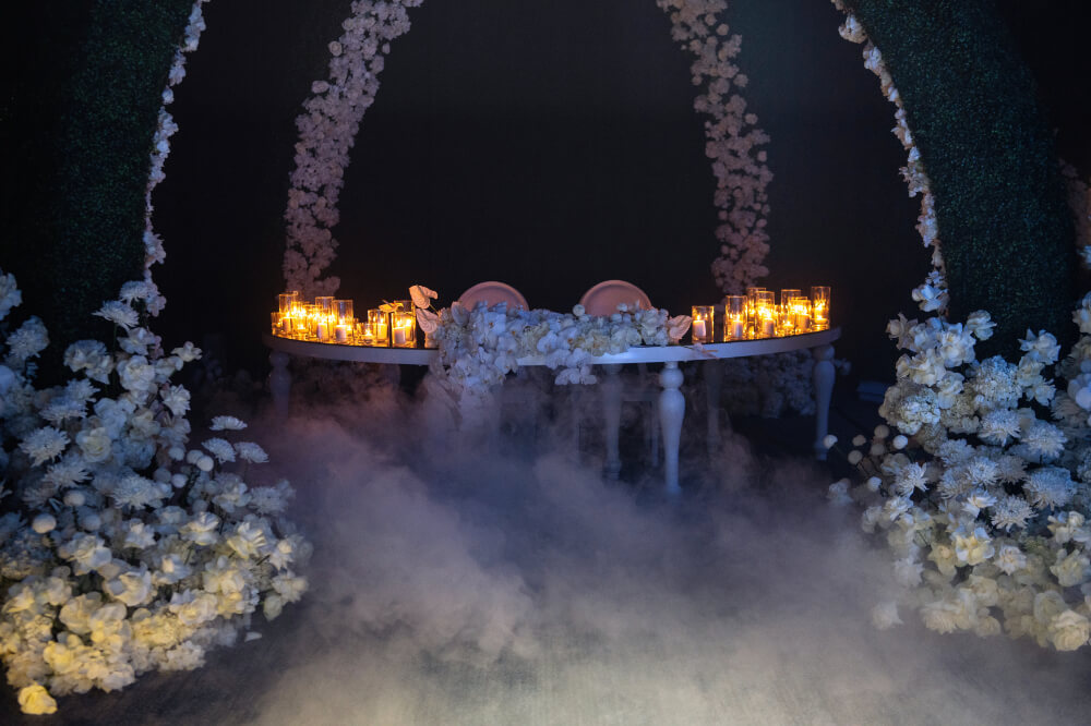 Romantic wedding table with candles, surrounded by white flowers and mist, creating an enchanting atmosphere.