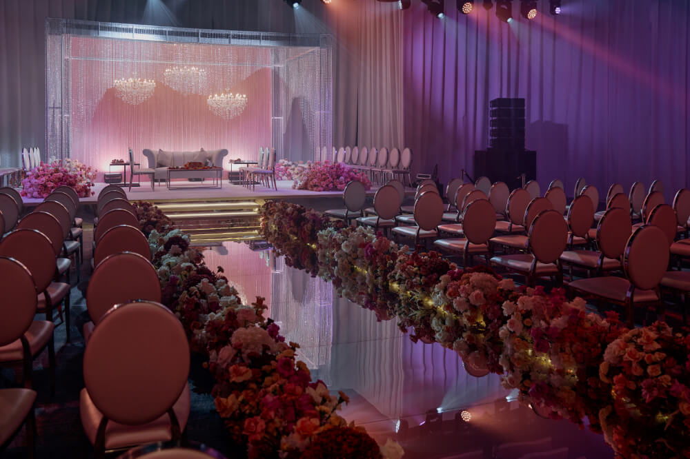 Elegant wedding venue with flower aisle, plush seating, chandeliers, and pink-lit stage for a romantic ceremony setup.