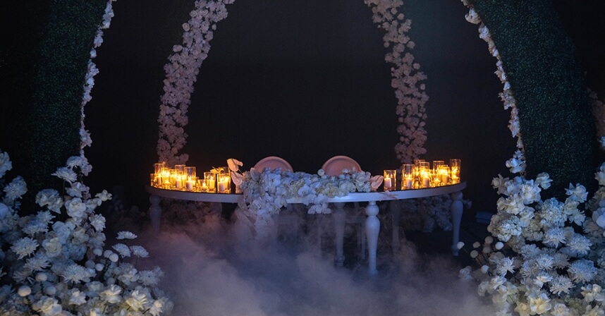 Elegant wedding table setting with candles and white flowers in a romantic, misty ambiance under floral arches.