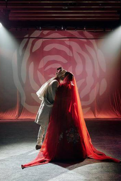A bride and groom embrace on a dimly lit stage with a red, floral-patterned backdrop, creating a romantic atmosphere.