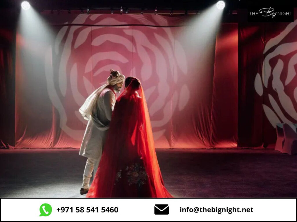 Bride and groom in traditional attire under spotlight at elegant wedding event, red floral backdrop, The Big Night.