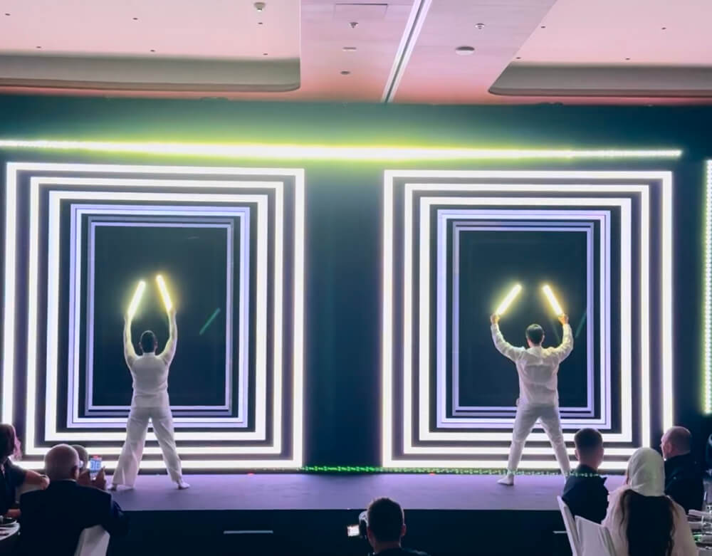 Performers with glow sticks on stage, surrounded by vibrant geometric light patterns, captivating audience at event.