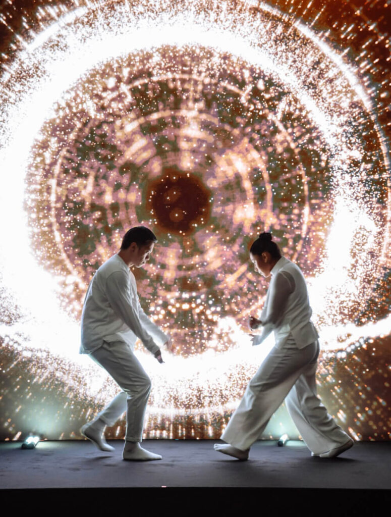 Two performers in white dance before a mesmerizing circular light display, creating a dynamic visual spectacle.