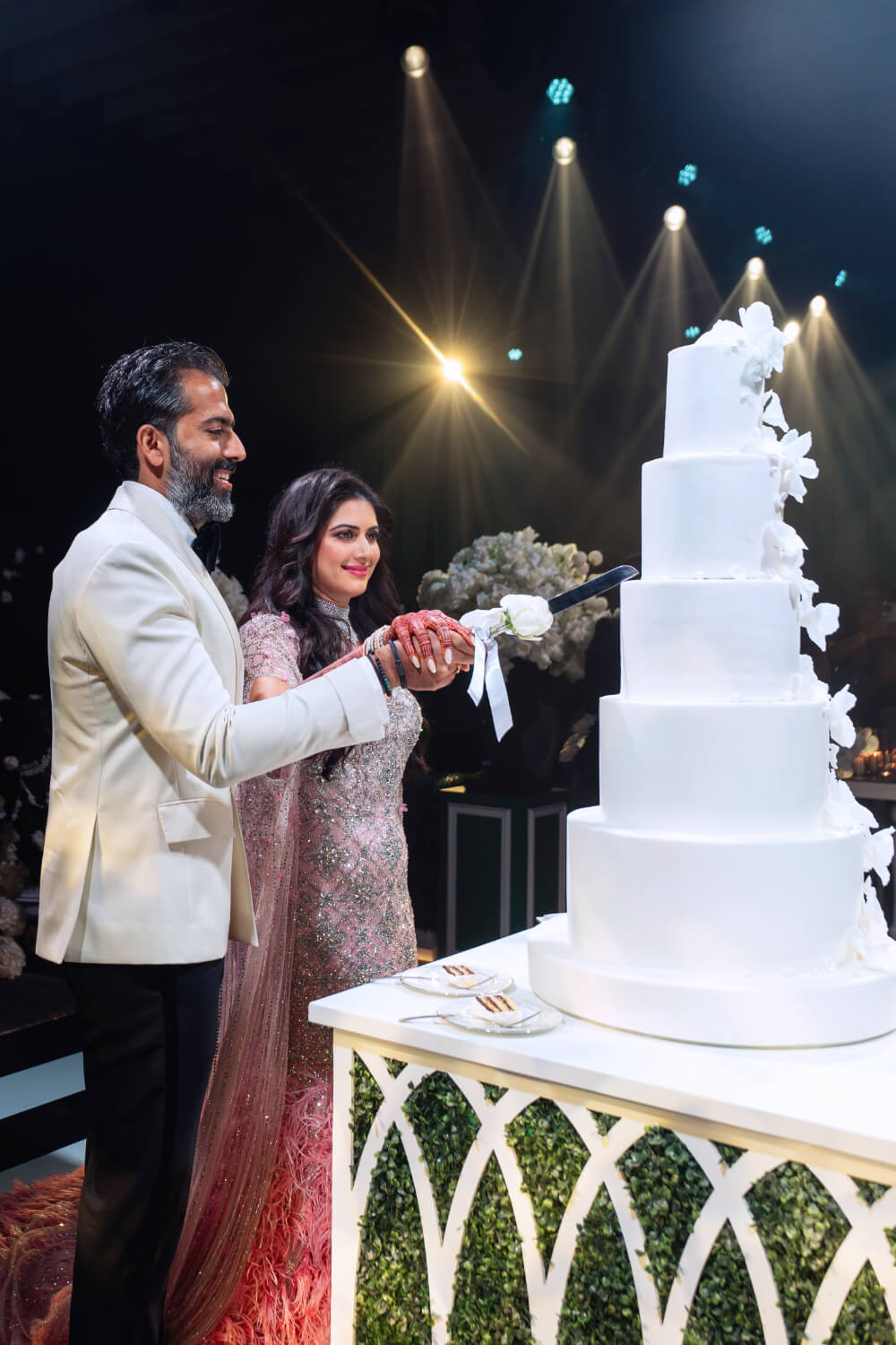 Couple cutting a large white wedding cake adorned with flowers at a reception, under celebratory lighting.