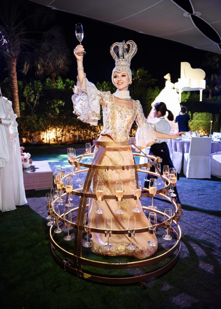 Woman in ornate dress with champagne tower at elegant outdoor event.