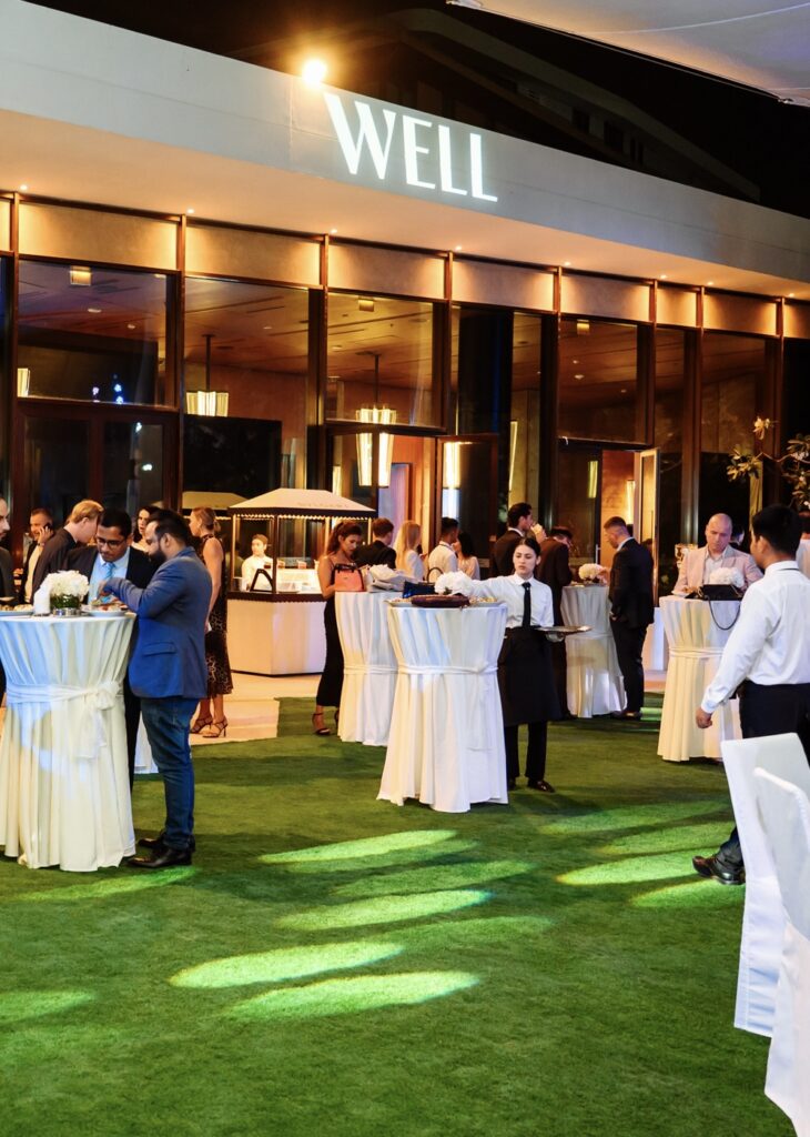 Outdoor event with guests networking by tables at night, warm lighting, and a venue sign reading WELL.