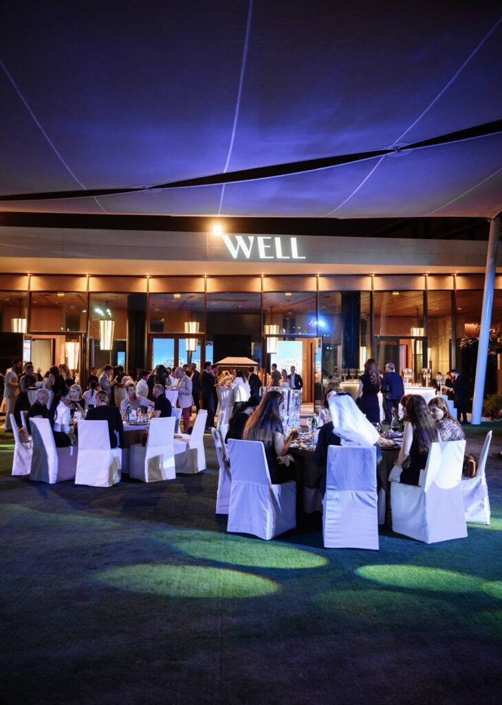 Outdoor evening event with guests seated at white-covered tables under illuminated venue canopy.