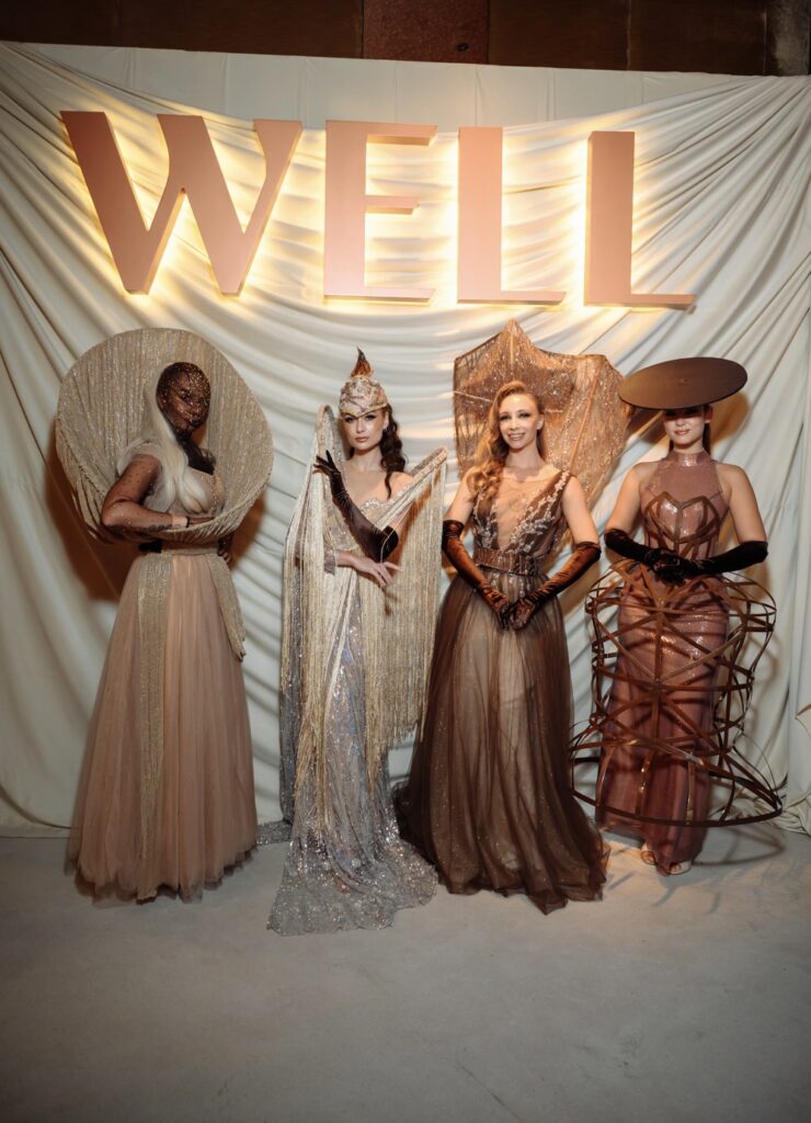 Four models in elaborate gowns at fashion event under WELL banner.