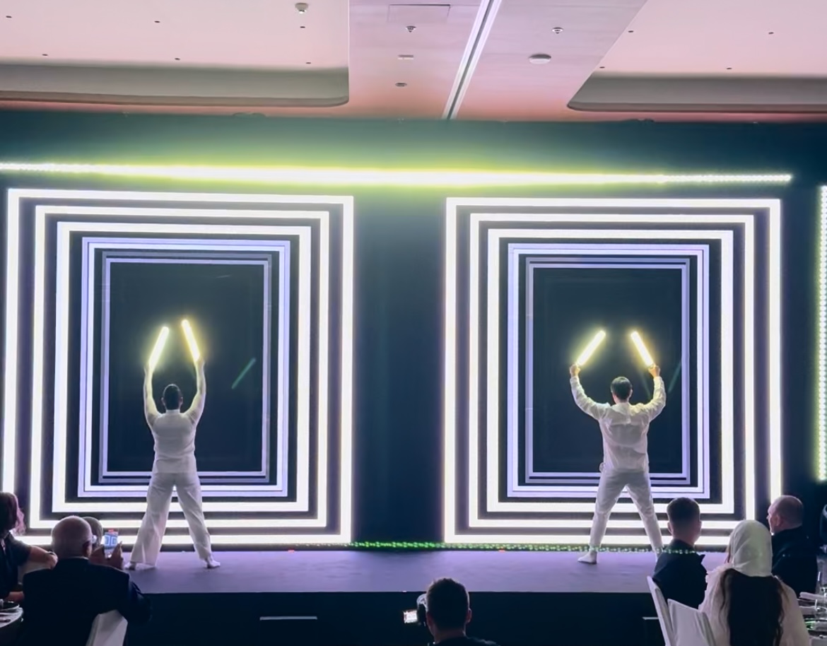 Performers in white outfits with glowing sticks in a light display, entertaining an audience at an indoor event.