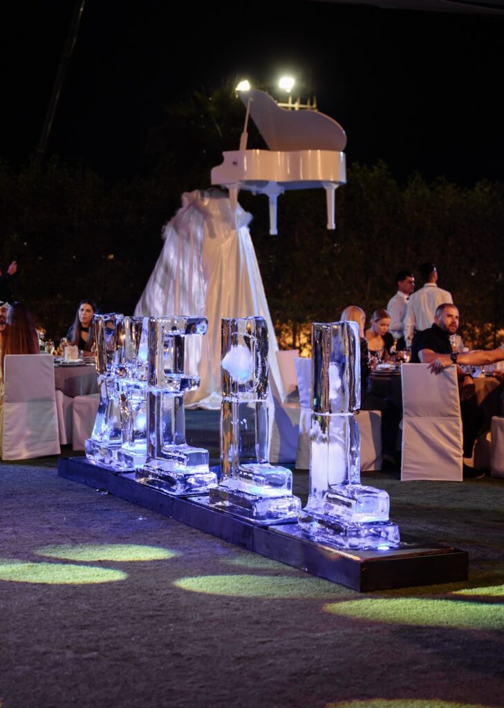 Ice sculpture letters in a festive event setting with guests dining under a floating white piano at night.