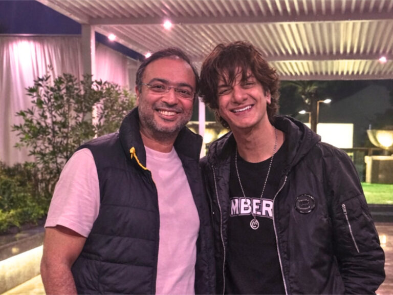 Two happy men smiling outdoors under a lit pergola.