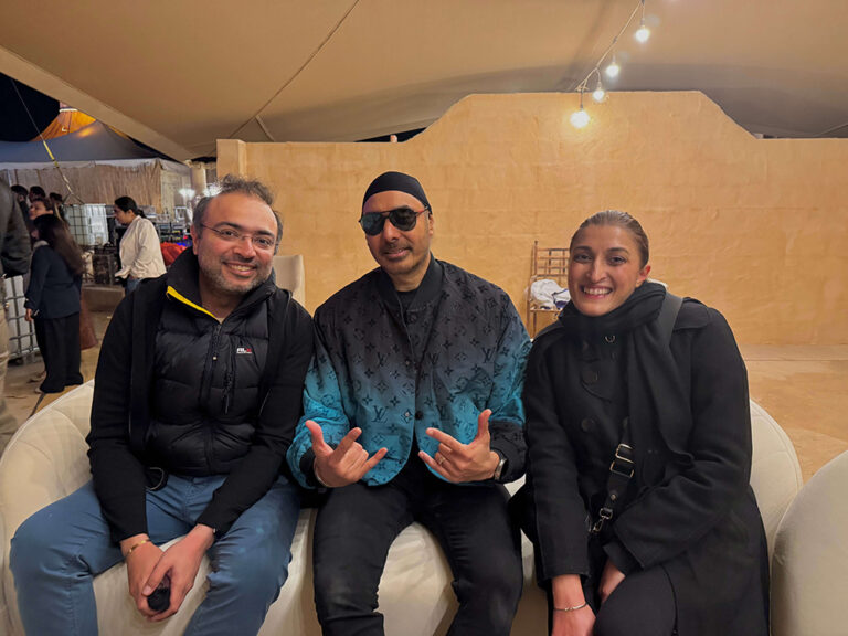 Group of three people smiling in a tented setting, seated on cushioned chairs.