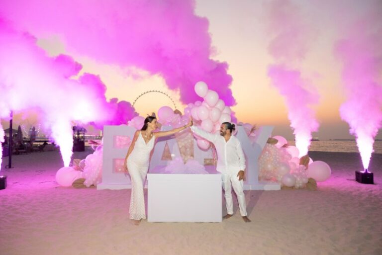 Couple celebrates gender reveal with pink backdrop and balloons on beach at sunset.