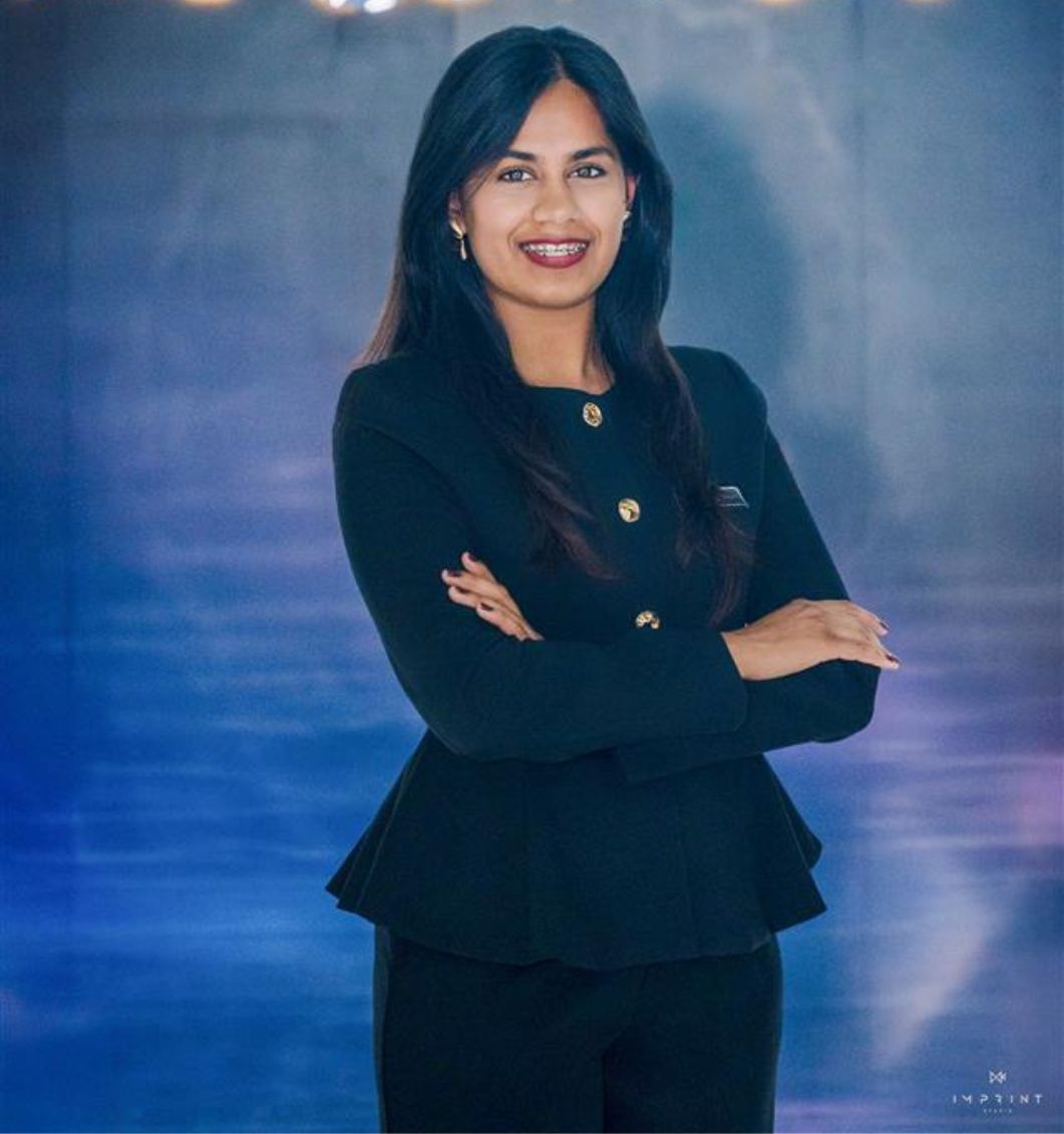 Confident woman in black suit smiling with arms crossed against a blue background.