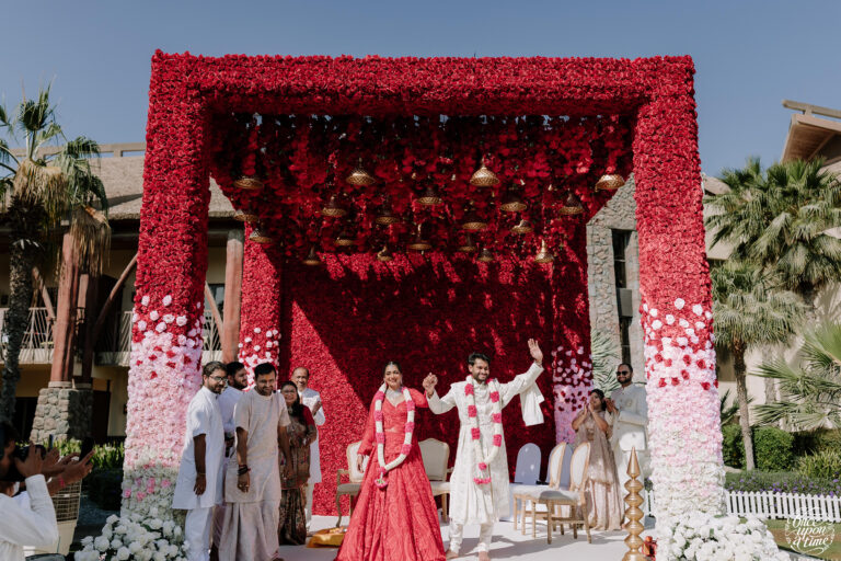 Bride and groom under a vibrant red floral wedding arch, celebrating in an outdoor ceremony.