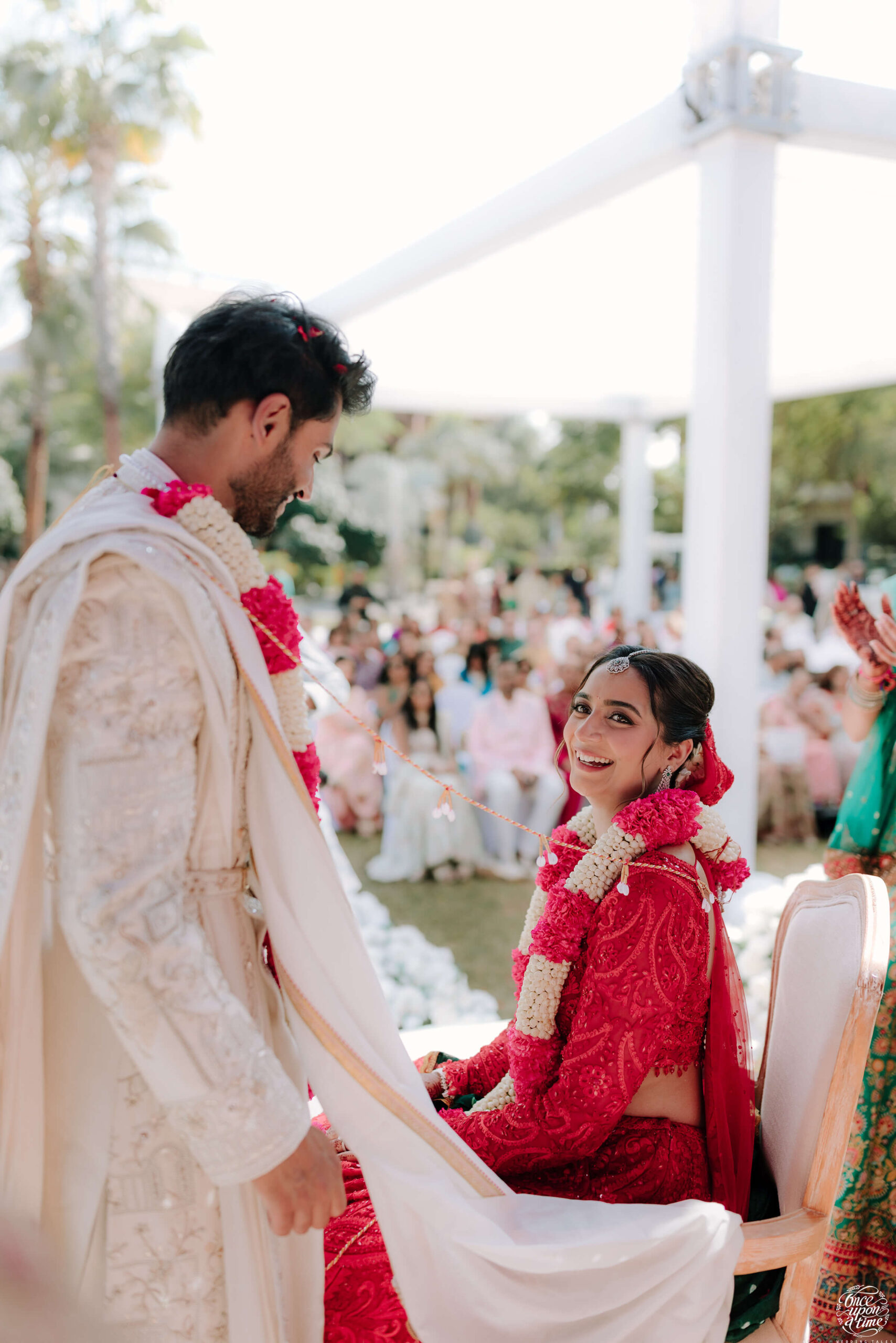 Bride and groom smiling during vibrant outdoor wedding ceremony, adorned in traditional attire with floral garlands.