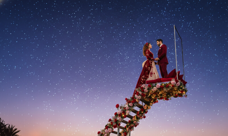 Couple in red attire on floral staircase under a starry sky at dusk.