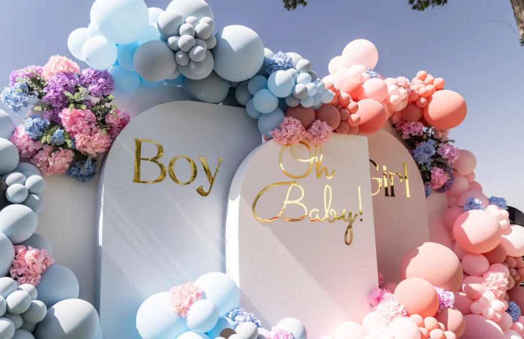 Gender reveal decorations with Oh Baby, Boy, and Girl signs surrounded by blue and pink balloons and flowers.