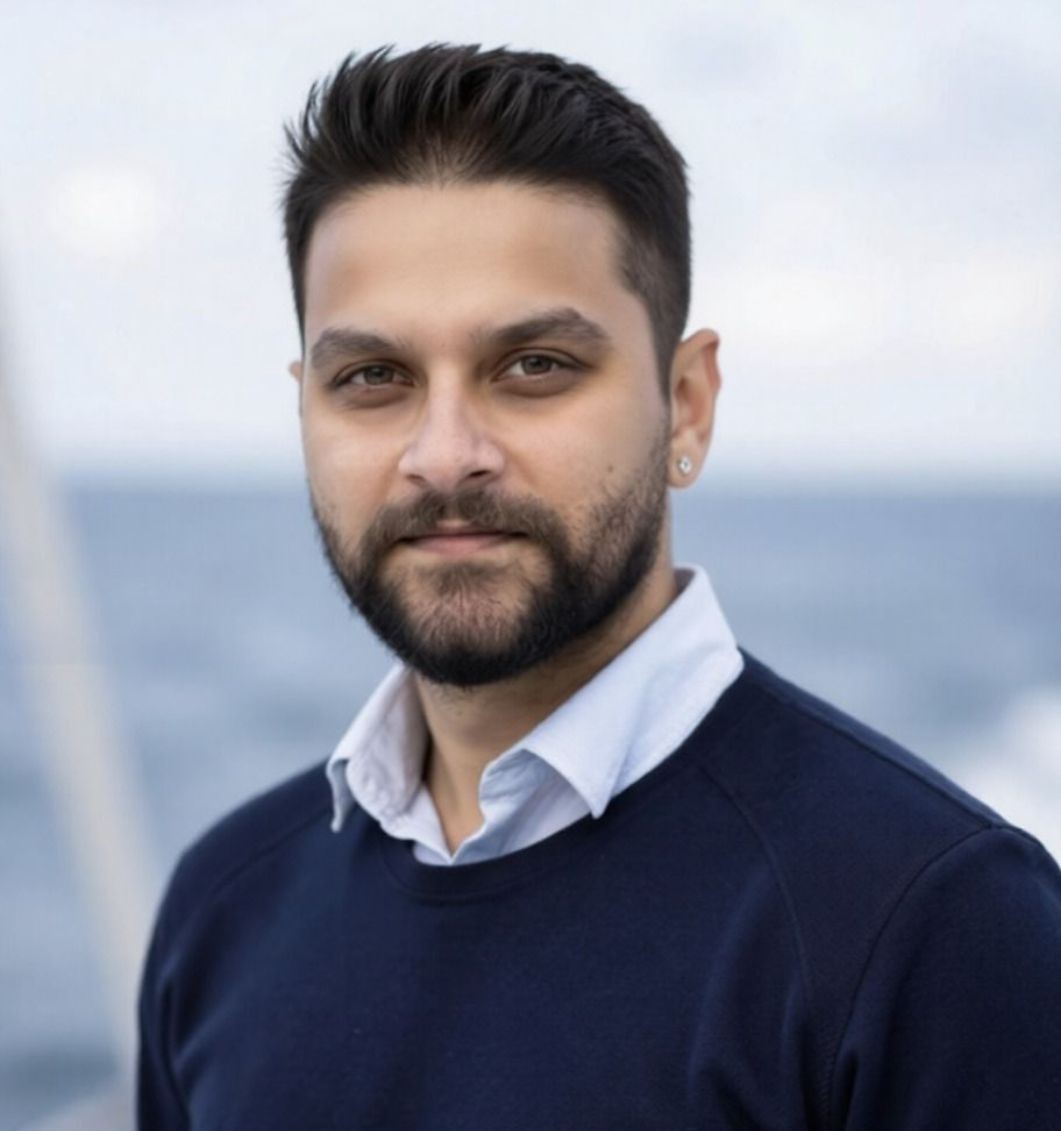 Professional man with a beard standing outside, wearing a navy sweater and white shirt, with a blurred ocean background.
