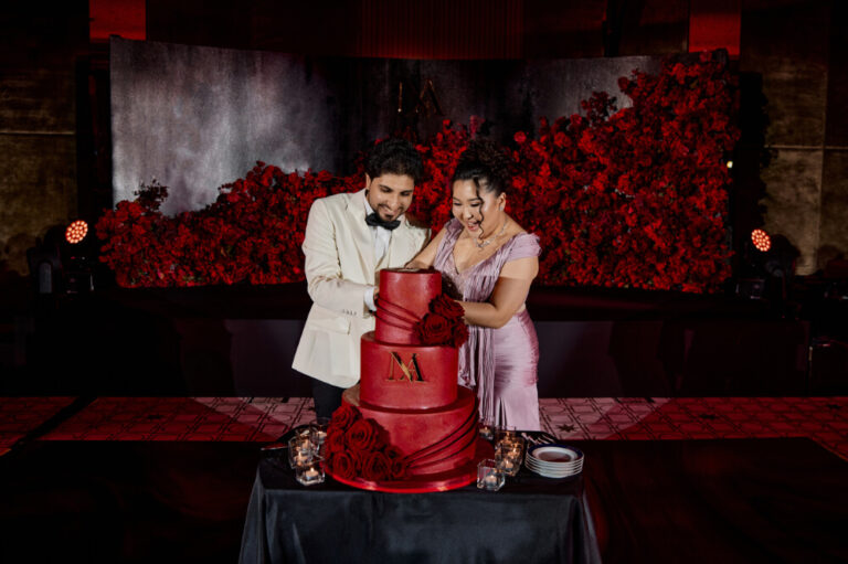 Couple cutting a red wedding cake adorned with roses and a monogram M in an elegant setting with floral backdrop.