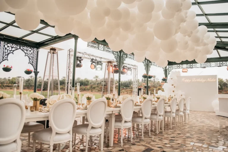 Elegant outdoor birthday setup with white balloons and decorated table under glass canopy.