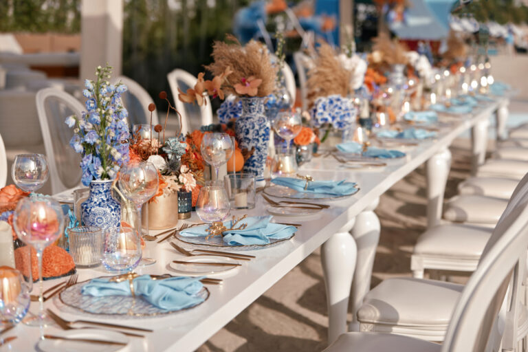 Elegant beachside table setting with blue napkins, floral centerpieces in vases, and candles for an outdoor event.
